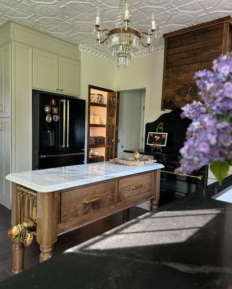 Our Modern Victorian kitchen stills📸 I hope y’all can appreciate the details in the space…the @americantinceilings tiles, the Nero Mist granite counters and tall backsplash with an ogee edge from @graniteworksstone , the antique Italian chandelier, the gorgeous custom dairy table island made by @clevelandcarpentry , the antique floral oil paintings I’ve been collecting , the 1800’s bed frame turned hood vent , the glass bifold pantry with brass @signaturehw Cremone bolts—it all came together ... Brass Hardware Kitchen, Bifold Pantry, Dairy Table, Bridgerton Love, Modern Victorian Kitchen, Modern Vintage Kitchen, Kitchen Cabinets Countertops, Countertops Marble, Kitchen Pantry Doors