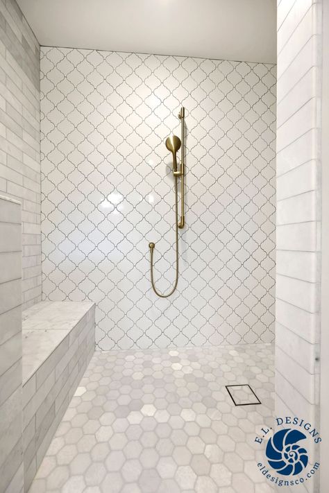 The image looks into a white open shower space. There is subway tile on the walls with an accent wall of glazed porcelain arabesque tile straight ahead. The shower floor is a marble hexagon tile, and the drain is a tile-in drain. The plumbing fixtures are brass and features a sitting bench on the left. White Subway Tile Shower With Marble Hexagon Floor, Arabesque Tile Shower Wall, Arabesque Tile Shower, Arabesque Tile Bathroom, Hexagon Bathroom, Arabesque Tiles, White Subway Tile Shower, Marble Subway Tile, Whole House Remodel