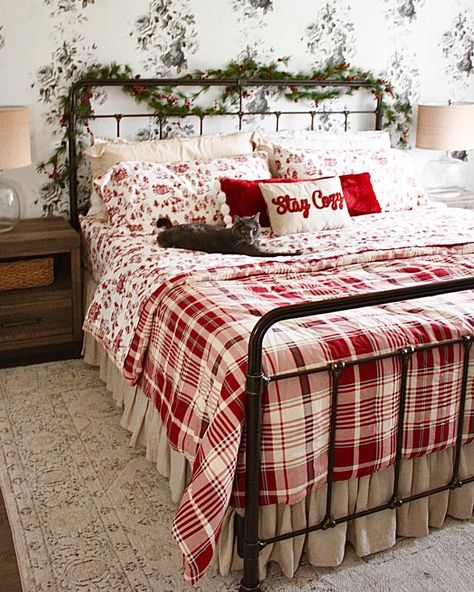 A black metal bed is pushed against a wall covered in white wallpaper with flowers on it. A garland is draped over the headboard. The bed is made with red and white patterned Christmas sheets underneath a red plaid comforter. Red and white throw pillows are placed on the bed that sits on a white area rug. Christmas Bedding Set Farmhouse, Christmas Bedding Ideas, Christmas Bedrooms, Red Comforter, Christmas Sheets, Holiday Bed, White Wall Bedroom, Christmas Bedding Set, Holiday Bedroom