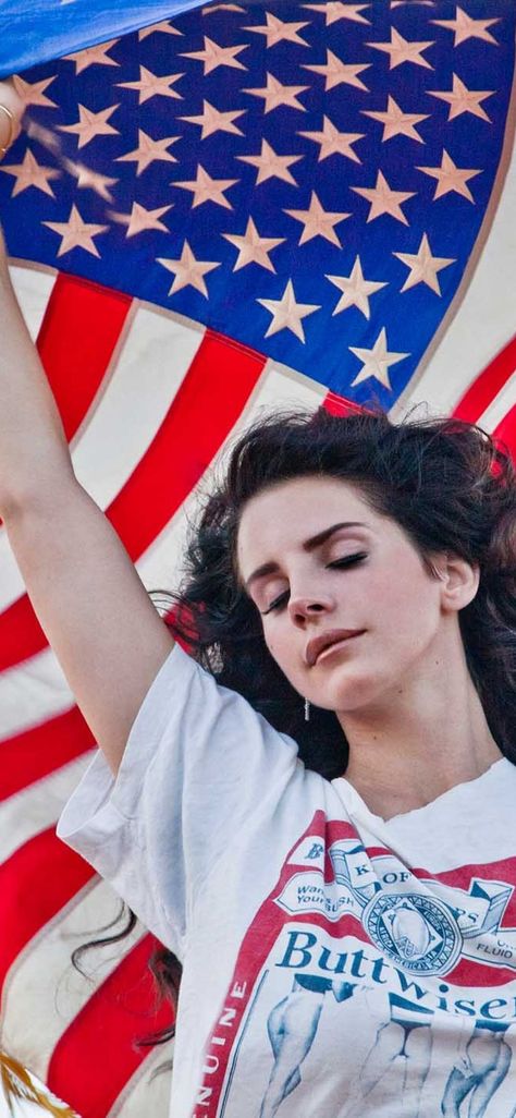 A white brunette, singer/songwriter Lana Del Rey,  stands with her eyes closed and wind blowing through her hair. She is holding the American flag above herself, letting it flow in the wind, and wearing a shirt that says Buttwiser, a spoof of Budweiser beer. Ride Lana Del Rey, Lana Del Rey Ride, Lana Del Rey Quotes, Iphone Wallpaper Music, Lana Del Rey Love, Lana Del Rey Lyrics, Brooklyn Baby, Americana Fashion, Vintage Americana