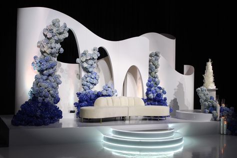 An ethereal white stage adorned with blue and white hydrangeas, a modern curved backdrop, and a white velvet love seat, creating a luxurious ambiance for a wedding reception. Modern Wedding Stage Design, Reception Stage Decoration Backdrops, Modern Wedding Backdrop, Modern Arches, Blue And White Hydrangea, Reception Backdrop, Wedding Stage Design, Wedding Backdrop Design, White Sofa