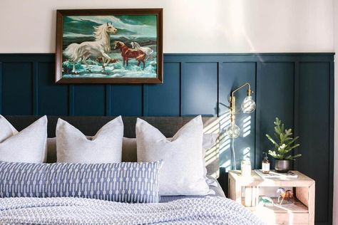 A dark wood-framed art piece is mounted above this gray upholstered bed clad in white and blue bedding. The bed is accompanied by a light wood bedside table lit by a gold and glass wall sconce. Add depth to the room with navy blue board and batten paneling behind the bed. Wainscotting In Bedroom, Blue Board And Batten Wall, Blue Board And Batten, White And Blue Bedding, Beige Living Room Walls, Gray Upholstered Bed, Bed Image, Wall Paneling Ideas, Light Wood Bed