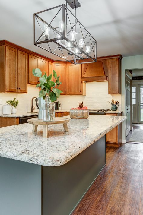 The contrasting warm and cool elements in this kitchen draw you in and makes you want to stay awhile. With white cabinets being so popular, these clients decided to go a different direction with brown maple wood wall cabinets. Adding a green island, light countertops, a white backsplash, and dark accents creates multiple textures, giving this kitchen dimension. The wall cabinets extend all the way to the large window, drawing in natural light, and the island offers bar seating. Island With Bar Seating, Island With Bar, Green Kitchen Island, Honey Oak Cabinets, Light Wood Kitchens, Kitchen Island Cabinets, Maple Kitchen Cabinets, Brown Kitchen Cabinets, Light Wood Cabinets