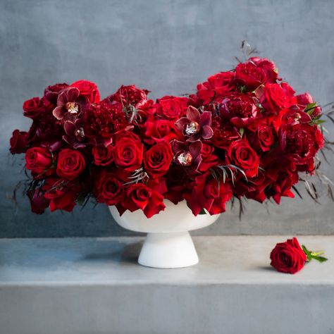 Sizes Available Explore this breathtaking floral arrangement showcasing opulent ruby red tones. Opt for the medium size with 70 lavish roses or go grand with the large size boasting 100 roses. The arrangement is adorned with abundant red roses, interspersed with deep burgundy peonies, and enhanced by graceful brown cymbidiums for an enchanting touch. Delivered in an immaculate white pedestal vase, this composition is a visual delight. Three sided arrangement. Red Peony Arrangement, All Red Floral Arrangements, Red And Gold Flower Arrangements, Red Floral Arrangements Modern, Large Rose Arrangements, All Red Flower Arrangements, Red White Floral Arrangements, Red Flower Arrangements Table, Red Flowers For Wedding