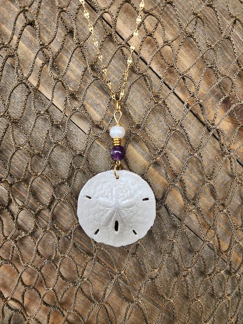 "Gorgeous, natural, handmade sand dollar necklace with genuine amethyst and freshwater pearl beads. The sand dollar is real and is painted in iridescent white pearl. It is accented with a real amethyst beads and  a freshwater pearl.  Amethyst is known for its calming power, so with this set you get the best of both worlds, a beautiful token from the sea and a gorgeous piece of the earth to compliment it. The pendant (approximately 1.5\" across) is set on an 18\", anti-tarnish stainless steel gol Sand Dollar Jewelry, Handmade Shell Necklace, Sand Dollar Craft, Sand Dollar Ornament, Sand Dollar Necklace, Pearl Amethyst, Sand Dollar Pendant, Surf Jewelry, Diy Jewelry Display