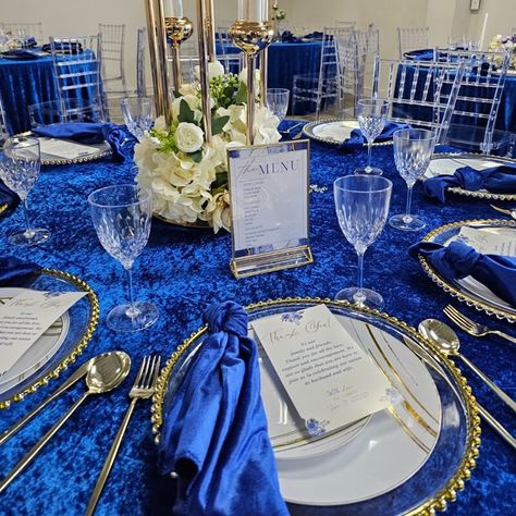 Behold the enchanting wedding banquet, resplendent in all its glory! The regal blue velvet tablecloth drapes gracefully, captivating hearts at first sight. Nestled upon clear round charger plates, royal blue satin dinner napkins exude opulence. Delicate white wax candles flicker inside long, transparent candleholders, casting a romantic glow. A small bouquet of faux white silk roses graces each table, adding a touch of everlasting beauty. Blue Velvet Tablecloth Wedding, Royal Blue Centerpieces Quinceanera, Royal Blue Centerpieces, Centerpieces Quinceanera, Velvet Tablecloth, Event Decor Ideas, Party Decorations Table, Group Dinner, Candles Flowers