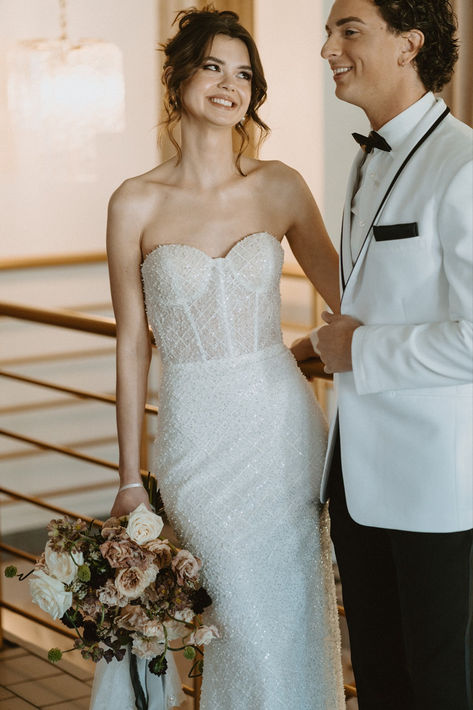 bride and groom smiling at each other. She is wearing a strapless wedding dress with a sparkle sequin fabric and is holding a white and beige floral bouquet. He is wearing a white suit with black pants and a black bow tie. Revelle Bridal, Wedding Dress Made With Love, Retro Bride, Bride Wedding Dress, Brides Wedding Dress, Bridal Shop, Bride Wedding, Bridal Boutique, Retro Vibe