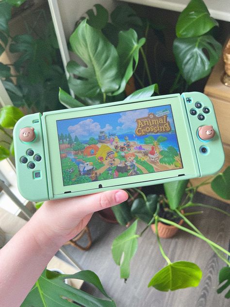 One hand holding a Nintendo Switch, green plants in the background. Animal crossing is on the switch screen. The Nintendo Switch had a matcha green soft case with cute bear thumb-sticks. Nintendo Switch Aesthetic Green, Nintendo Switch Covers, Green Nintendo Switch, Aesthetic Nintendo Switch, Aesthetic Nintendo, Nintendo Aesthetic, Green Pc, Switch Case, Nintendo Switch Case