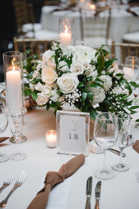 Low, lush, and classic white and green wedding centerpiece with candles to surround. Modern mocha tied napkins at each place setting. Acrylic table number frame. PC: Asya Photography https://asyaphotography.com/ Wedding Centerpieces White Flowers And Greenery, Central Piece Wedding Table, White Floral Centerpieces Round Table, White And Greenery Centerpiece, Wedding Flowers White And Green Centerpieces, White And Green Wedding Table, Green And White Wedding Centerpieces Round Table, White And Green Wedding Table Decor, Low Wedding Centerpieces Round Table