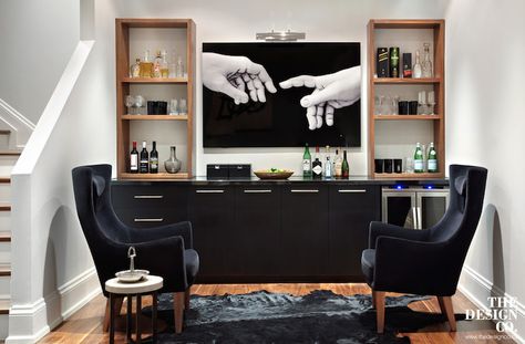 Contemporary basement with wet bar boasting black base cabinets adorned with chrome pulls and fitted with two-door glass-front beverage fridge topped with sleek black countertops topped with walnut stained shelving units filled with wine glasses and libations flanking a flatscreen TV attached to the wall illuminated by a picture light. Beautiful Basements, Rustic Basement Bar, Basement Bar Plans, Contemporary Basement, Basement Bar Design, Basement Bar Designs, Game Room Basement, Game Room Bar, Black Countertops