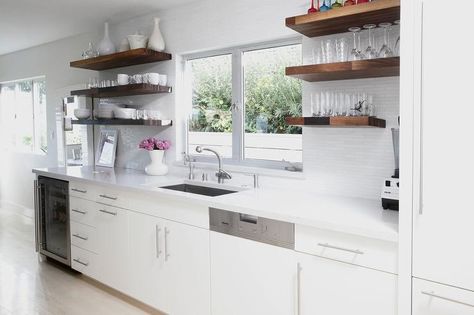 Chic kitchen features white flat front cabinets paired with white quartz countertops fitted with a sink and satin nickel faucet under a window flanked by stacked wood shelves lining a glossy white tiled backsplash. Shelves Between Windows, White Kitchen Floating Shelves, White Kitchen Open Shelves, Small Kitchen Cabinet Storage, Flat Front Cabinets, Wooden Shelves Kitchen, Wood Shelves Kitchen, Floating Kitchen Shelves, Small White Kitchens