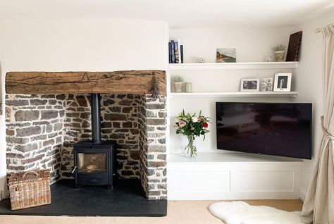 Black wood burning stove is placed inside a stone fireplace with black hearth and rustic wood mantel. A flat panel TV is positioned inside a white built-in media unit next to the fireplace. White Fireplace Mantels, Painted Stone Fireplace, Grey Stone Fireplace, White Stone Fireplaces, Woodburning Stove Fireplace, Fireplace Facing, Fireplace Feature Wall, Stacked Stone Fireplaces, Patterned Tile Backsplash