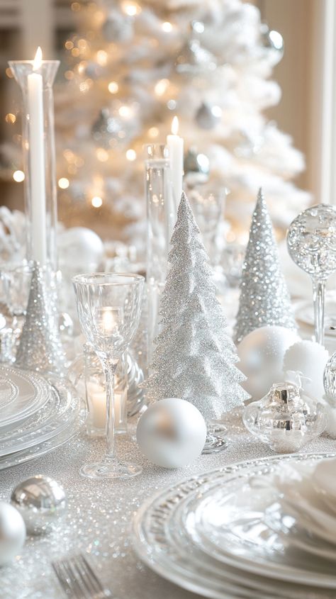 Elegant white and silver Christmas table setting with crystal glasses, candles, and glass tree figurines in a modern design. White And Silver Christmas Table Setting, All White Christmas Party Decorations, New Years Dining Table Decor, White And Silver Table Settings, White Christmas Dinner Party, Winter Wonderland Tablescape Ideas, Silver And White Christmas Table Decor, White Table Setting Christmas, Snowy Tablescape