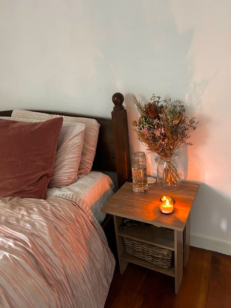 A bedside table with dried pink flowers, glass of water and a candle burning. The flowers match the bedding and pillow colours. Clean Cosy Bedroom, Nightstand Decor Aesthetic, Cozy Nightstand, Night Stand Aesthetic, Aesthetic Nightstand, Nightstand Bedroom, Warm Bedroom, Bedroom Nightstand, Nightstand Decor