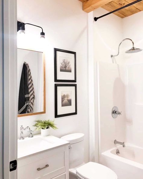 A chrome faucet and shower head add a modern touch to a bathroom with a wood ceiling. White walls are accessorized by black picture frames and vanity lighting. The wood-framed mirror is affixed above a small white vanity with a chrome faucet. Small White Vanity, White Bathroom Walls, Grid Shower Door, White And Chrome Bathroom, Shiplap Bathroom Wall, Bathroom Lighting Ideas, White Vessel Sink, Shiplap Bathroom, Black Floor Tiles