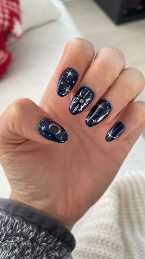 Navy shimmer nail polish with gold stars of different sizes and shapes, a large sun on the middle finger and a crescent moon on the thumb. Inspired by the book: A Court of Frost and Starlight Dark Blue Moon And Star Nails, Manon Blackbeak Nails, Simple Ravenclaw Nails, Witchy Celestial Nails, North Star Nail Design, Night Theme Nails, Star Theme Nails, Star And Glitter Nails, Loki Themed Nails