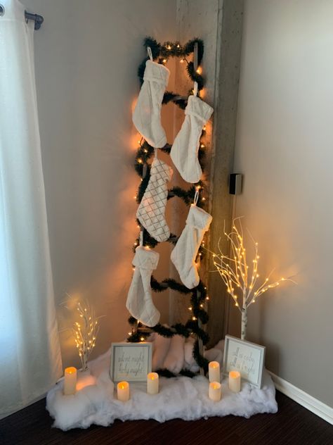 A ladder in the corner of an apartment, covered in garland and Christmas lights. Five white stockings are tacked on the ladder. The bottom of the ladder is surrounded by fake snow, light up birch trees, and holiday themed signs. Stocking With No Fireplace, Stalking Ideas For Christmas, Stocking Placement Without Fireplace, Christmas Stockings Without Fireplace, Stocking Organization, Creative Ways To Hang Stockings Without Mantle, Stocking Without Fireplace, Stockings No Fireplace Ideas, Christmas Stockings No Fireplace