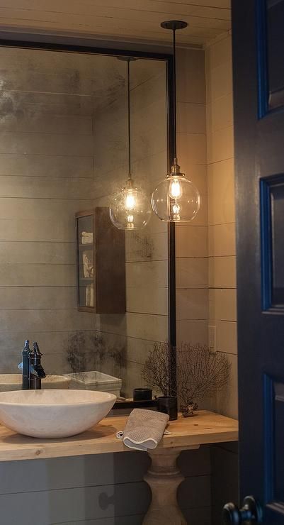 A black door opens to a gray powder room features walls clad in gray shiplap lined with a blond wood balustrade vanity topped with a marble bowl sink and bronze faucet under a black framed mirror lit by a single glass globe pendant. Powder Room Pendant Lights Over Vanity, Hanging Vanity Lights Bathroom, Hanging Light In Powder Room, Ensuite Pendant Lighting, Hanging Bathroom Pendant Lights, Wall Lights In Bathroom, Oversized Mirror Powder Room, Pendant Light In Front Of Mirror, Powder Bathroom Pendant Lighting