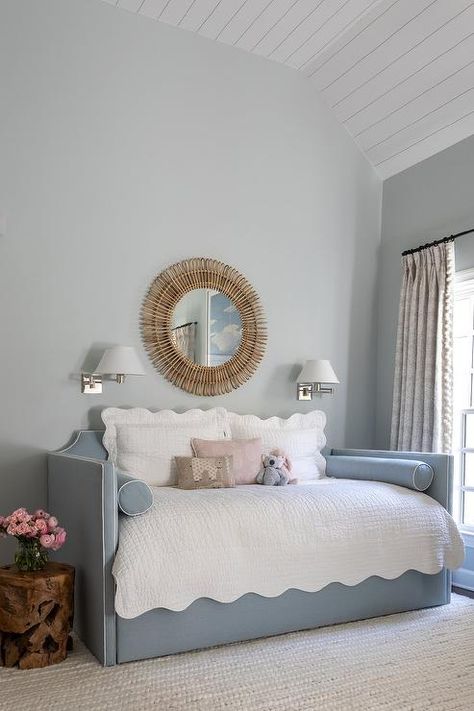 Beneath a vaulted plank ceiling, a blue French daybed is accented with white scalloped bedding and blue bolster pillows lit by polished nickel swing arm sconces fixed on either side of a rattan sunburst mirror. Nursery Accent Table, Scalloped Bedding, Nursery Daybed, Gray Upholstered Bed, Daybed Ideas, Transitional Nursery, Daybed Pillows, White Daybed, Bold Bedding