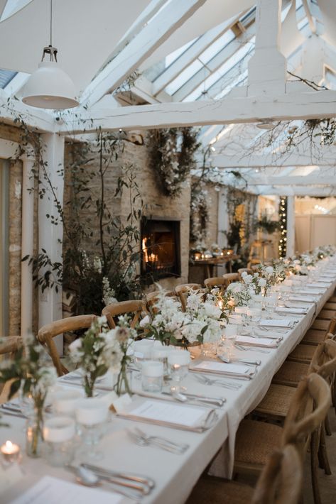 A white and green theme of a winter wedding. There is a long table covered in a linen cloth, wooden chairs, and a fire place glows in the background. There are rustic white wooden beams above. The table is decorated in green foliage and white flowers and candles. Cotswold Wedding, Uk Countryside Wedding, Cotswolds Wedding Venues, Uk Farm Wedding, Daylesford Farm, Woodland Wedding Venues Uk, Wedding Venue Scotland, Winter In Australia, Winter Wedding Venues