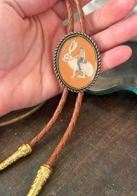 This awesome bolo tie has a cowboy riding a jackrabbit print with a clear resin coating and metal backing. Handmade in our shop! The cord is brown and made of genuine woven leather. Our bolos pair nicely with many of our belt buckles! They make wonderful gifts.    The western bolo tie rope length is 39' ;  pendant size is 1 1/2'' x 1 1/4'' Western Bolo Tie, Country Rings, Jack Rabbit, Bolo Ties, Mens Necklace, Tie Gifts, Bolo Tie, Resin Coating, Cow Boy