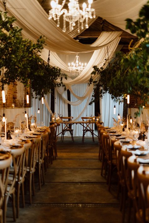 This is showing a barn wedding dining area. The overall vibes are rustic luxury. On the ceiling are glass chandeliers, white drapes hanging across the ceiling, directly above the wooden long dining tables are hanging boards with ivy on them. On the tables are white table runners, vases, candlesticks, tableware and glassware. In the middle of the two rows of dining tables is a sweet heart table at the end. Wooden Crossback Chairs Wedding, Low Ceiling Draping Wedding, Wedding Ceiling Drapery, Wedding Decor Hanging From Ceiling, Wedding Ceiling Decorations Draping, Curtain Ceiling Design, Wedding Drapes Ceiling, Black Draping Wedding, Wedding Decor Draping