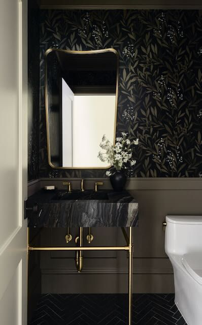 Elegant small bathroom featuring a dark leafy wallpaper that adds depth, complemented by a sleek black marble vanity with gold accents, a sophisticated wall-mounted mirror, and a minimalist white toilet, creating a luxurious yet intimate ambiance with rich textures and contrasting colors. Moody Small Bathroom, Black Marble Vanity, Larchmont Ny, Nyc Home, White Toilet, Marble Vanity, Small Bathroom Decor, Wall Mounted Mirror, Textured Wall