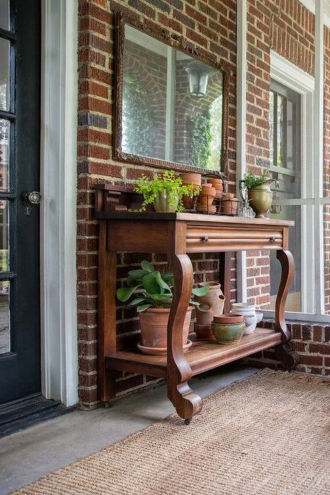 Sunroom features red brick walls, a console table under a mirror and a fringe jute rug. Brick Sunroom, Bungalow Attic, Sunroom Extension, Red Brick Pavers, Vintage Bungalow, Brick Accent Wall, Brick Hearth, Wicker Lounge Chair, Red Brick Walls