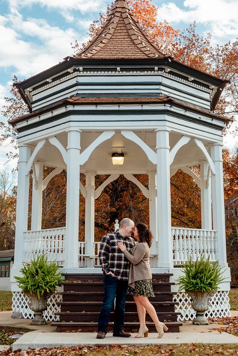 A heterosexual couple stands under a gazebo, foreheads pressed together and looking into each other's eyes. The sky behind them is blue with a few clouds, and the leaves are bright orange. Gazebo Couple Photoshoot, Gazebo Couple Pictures, Engagement Photos Gazebo, Gazebo Engagement Pictures, Engagement Photos Town, Small Town Couple Photoshoot, Gazebo Photoshoot Ideas, Small Town Engagement Photos, Small Town Photoshoot