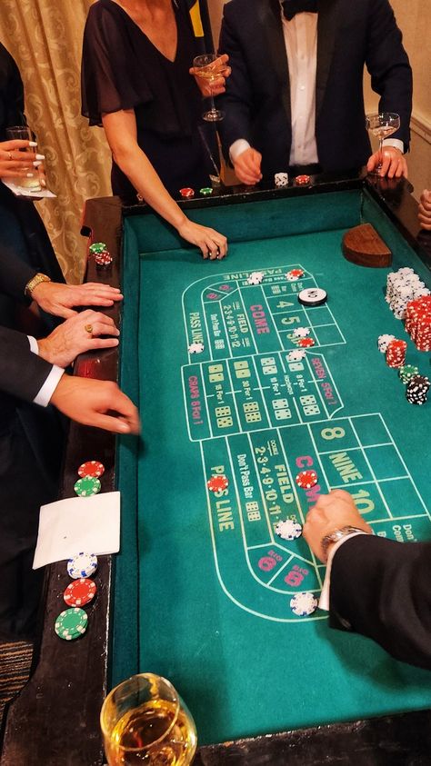 A craps table surrounded by tux covered hands, wine glasses, ladies in evening gowns. Wine glass in the lower corner, dice and chips on the table as play is in session Craps Table, Imaginative Writing, Casino Room, Casino Table Games, Surprise 60th, Casino Jackpot, Green Room Decor, Casino Party Games, Casino Birthday