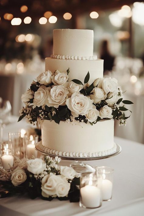 Three-tier white wedding cake adorned with white roses and surrounded by candles. Get enchanted by wedding decor ideas that remain elegant across the ages while impressing even the pickiest Aunt Mildred. White Tiered Wedding Cake, Wedding Cake Simple White, Wedding Cake Table Decor, Timeless Wedding Decor, Wedding Cake Table Decorations, Floral Archway, Dream Wedding Cake, Wedding Decor Ideas, Cake Table Decorations