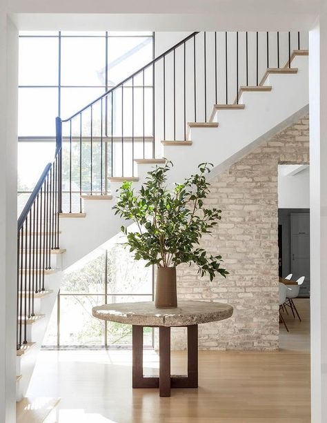 Minimalist foyer boasts a round concrete table topped with a potted plant and positioned in front of white staircase finished with an iron handrail and spindles and wood treads illuminated by natural light from a large black factory window accented with wall half covered in brick. Boho Floral Nursery, Staircase Window, Foyer Staircase, Floral Nursery Decor, Mediterranean Style Home, Interior Finishes, Open Staircase, Mediterranean Home Decor, Take The Stairs