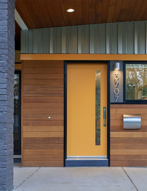 A cheery yellow front door with a tall off-center window slit is the central attraction, while the midcentury modern silver house numbers, door handle, and mailbox provide stylish support. The walls are clad in horizontal wood beams of a warm orangey tone. Above the door, vertical metal cladding reaches to the wood ceiling, repeated across the top until it disappears off the photo. Mid Century Front Door, Foyers And Entryways, Mid Century Modern Front Door, Mid Century Modern Entryway, Mid Century Modern Door, Orange Front Doors, Yellow Front Doors, Mid Century Modern Exterior, Entrance Halls