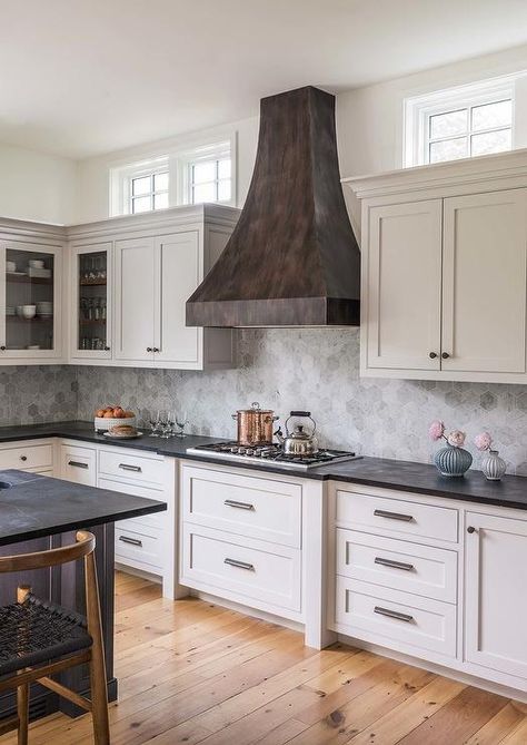 Windows are located above white shaker cabinets lined with crown molding and positioned beneath windows located on either side of a distressed black metal French range hood. French Range, Wood Floor Design, Recessed Panel Cabinets, Wood Floor Kitchen, Hood Design, White Shaker Cabinets, Kitchen Range Hood, Cottage Kitchens, Shaker Cabinets