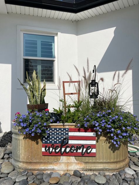 A variety of zone, sunlight, and soil plants inside a livestock tank with added visual interest such as a rustic bucket, a ceramic hen nestled in the plants, a small shepherd’s hook with an antique lantern, and a patriotic sign hanging on the front. At night, the display, which sits between the driveway and front door, is lit up by a solar spotlight. Repurposed Water Trough, Water Trough Ideas Backyards, Cow Trough Garden, Stock Tank Repurposed, Trough Flower Planters, Trough Flower Garden, Water Tank Planter Ideas, Metal Horse Trough Planters, Old Water Trough Ideas