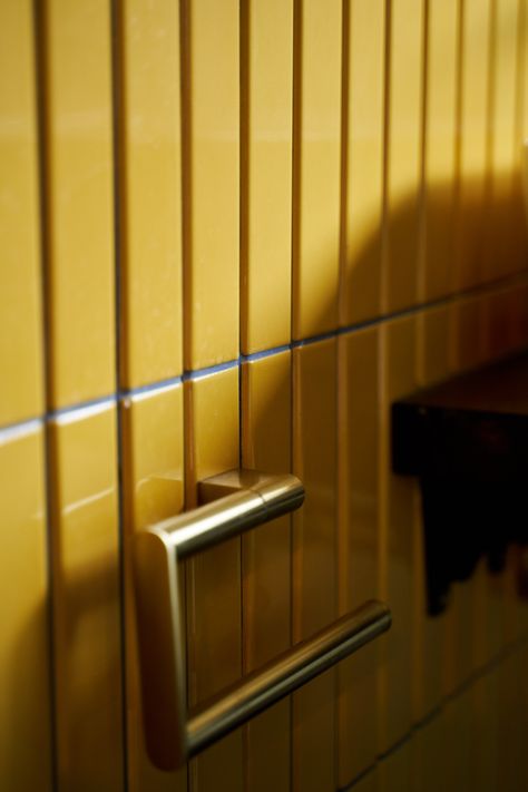 A close-up view of vibrant mustard yellow vertical tiles in a bathroom, paired with a sleek brushed brass towel rail. The tiles have a glossy finish, reflecting light and adding depth to the wall. The combination of bold color and luxurious brass accents creates a striking, contemporary look, perfect for adding warmth and style to a modern bathroom space. The shadows cast on the tiles enhance their textured appeal, making the brass pop against the rich yellow background. Mustard Yellow Bathroom, Chic Modern Bathroom, Small Family Bathroom, Family Bathroom Design, Yellow Bathroom Tiles, Unique Bathrooms, Brushed Brass Hardware, Chevron Tile, Traditional Bathrooms