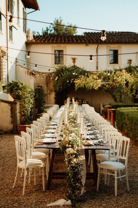 Wedding reception with a beautiful family style long wooden table, white romantic chairs, table runner made of eucalyptus, tea roses and white garden flowers, white taper candles in golden candelholders and vintage table settings under the string lights at a beautiful Italian garden of the 17th century Tuscan villa. Long Table Dinner, Charleston Elopement, Long Table Wedding, Southwest Wedding, Summer In Italy, Table Dinner, Wedding In Tuscany, Under The Tuscan Sun, Tuscan Wedding