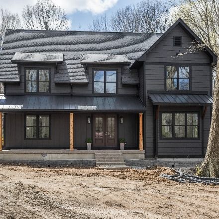 A charcoal farmhouse with black roof. Brown double doors with greenery and rug. Black light fixtures on sides of double doors. Photography by The Home Aesthetic & Builder Josh Loewen. Dark Gray Farmhouse, Black Gray House Exterior, Dark Gray Siding With Black Metal Roof, Charcoal Exterior House Colors Farmhouse, Charcoal House Exterior Black Trim, Black Modern Exterior House, Charcoal Gray Exterior House, Black House Gray Roof, House With Black Siding