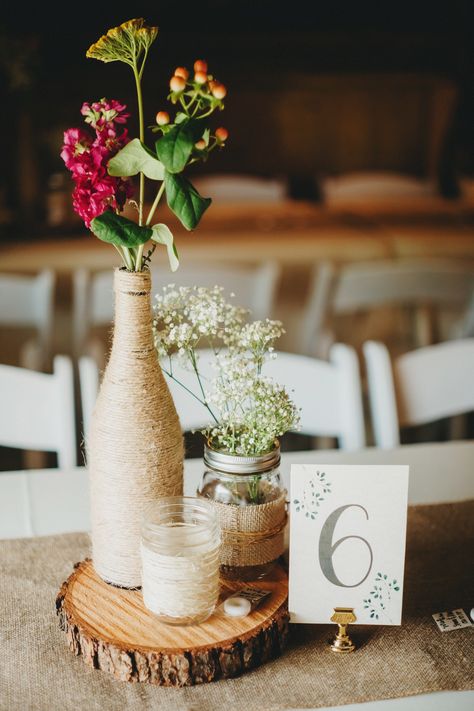 Rustic Center pieces, twine wrapped wine bottles, mason jars, paper cards, wood rounds  Rustic Grace Estate, Van Alstyne, TX  Photocred: Two Pair Photography Wedding Centerpiece With Wine Bottles, Wine Bottle Mason Jar Centerpiece, Rustic Wine Bottle Centerpieces, Boho Wine Bottle Centerpieces, Wine Bottle Centrepiece, Whiskey Bottle Wedding Centerpieces, Wine Bottle Diy Wedding, Winter Wedding Table Centerpieces, Wine Bottle Wedding Centerpieces