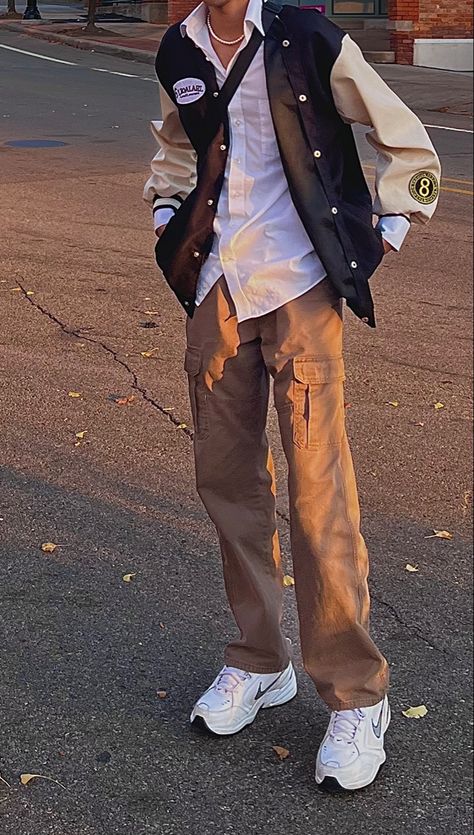 neck down picture of man wearing a black, tan, and white colored varsity jacket, with a white collared shirt on the inside and brown cargo pants. pearl necklace and nike shoes, with a crossbody small black bag. Beige Varsity Jacket Outfit Men, Brown Varsity Jacket Outfit Men, Beige Varsity Jacket Outfit, Brown Varsity Jacket Outfit, Black Varsity Jacket Outfit, Black And White Outfit For Men, Nike Varsity Jacket, Brown Cargos, Varsity Outfit