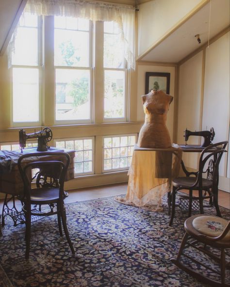 Soft light trickles in from floor-to-ceiling windows, illuminating two antique sewing machines on either side of the room, with a rough spun mannequin form on top of one of the tables. Sewing Room Aesthetic Vintage, Victorian Seamstress Aesthetic, Dark Academia Sewing Room, Victorian Dorm Room, Victorian Sewing Room, Sarah Winchester, Cottagecore Painting, Vintage Sewing Rooms, Victorian Sewing