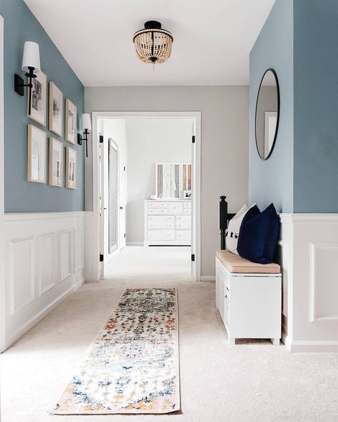 This hallway boasts beautiful color contrasts with white wainscoting fitted along a muted blue wall. A white bench seated under a black circle mirror creates a place to sit and admire a light wood frame gallery wall on the other side. Blue Walls With White Wainscoting, Decorating A Narrow Hallway, Modern Wainscoting Ideas, Hallway Mudroom, Blue Hallway, White Hallway, Hallway Paint, Hall Colour, Window Mudroom