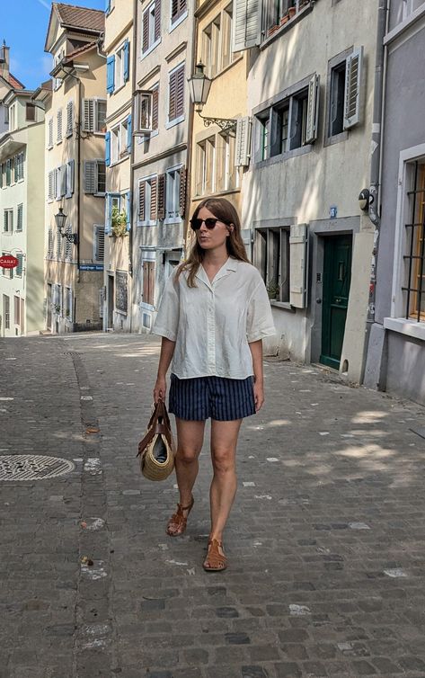 A woman stands on a cobbled street with colourful houses in the background. She wears a short-sleeved shirt with boxer shorts, leather fisherman sandals and a basket bag. Outfits In Switzerland, Outfits With Fisherman Sandals, Fisherman's Sandals Outfit, Fisherman Sandals Women Outfit, Fisherman Sandals Street Style, Fisherman Sandals With Socks, Fishermen Sandals Outfit, Angharad Jones, Leather Sandals Outfit