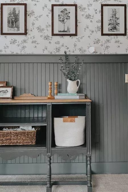 Farmhouse interior design style meets grand millennial design style in my dark moody living room. I love how grizzle gray by Sherwin-Williams brings so much dimension to my farmhouse living room while decorating with vintage ads the granny chic character that I love. This dark board and batten wall accent turned out so well. I added the floral wallpaper above for even more coziness. You can shop all of these home decor ideas on my LTK. Home Decor Ideas Wallpaper, Rustic Wainscoting Ideas Farmhouse Style, Board And Batten Around Thermostat, Floral Wallpaper With Wainscoting, Beadboard And Wallpaper Accent Wall, Black Board And Batten With Wallpaper, Gray Paneling Walls, Dark Grey Board And Batten Wall, Board Batten Living Room