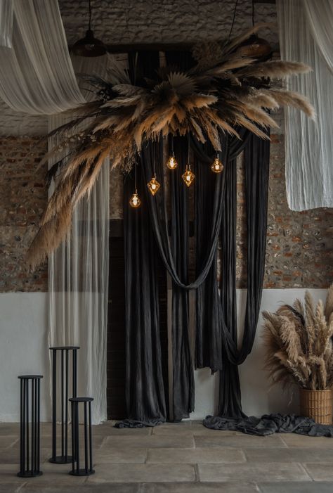 This image shows a barn wedding ceremony area. The main focus is on a hanging dried floral installation. The installation consists of normal cream pampas leaves and dark black ones, creating a depth in the installation. There are also uniquely shaped light bulbs hanging from the installation. Behind the installation are black and white drapes hanging across the venue ceiling and down the wall. Below the flowers are black plinths. Black Wedding Drapery, Black Wedding Draping, Suspended Floral Arrangements, Fabric Hanging From Ceiling, Backdrop Ideas For Wedding, Unique Backdrop Ideas, Chelsea Fabric, Wedding Draping Backdrop, Dried Flower Installation