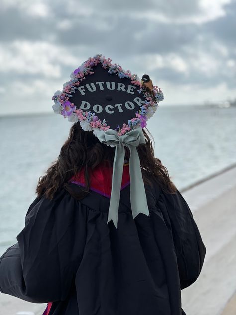 Picture of graduation cap with white roses on the bottom and pink, purple, and blue flowers on the top lining the cap, and surrounding the words “Future Doctor” in mint green in the center. A fake bird perches on the edge of the caps flowers. A large mint green bow in the same fabric and color shade as the words above is on the edge of the cap pointing towards her graduation gown Graduation Cap Designs Doctor, Graduation Cap Designs Medical School, Future Doctor Grad Cap, Future Doctor Graduation Cap, Graduation Cap Designs College Medical, Pre Med Graduation Cap, Medical Graduation Cap, Biology Graduation Cap, Graduation Cap Diy