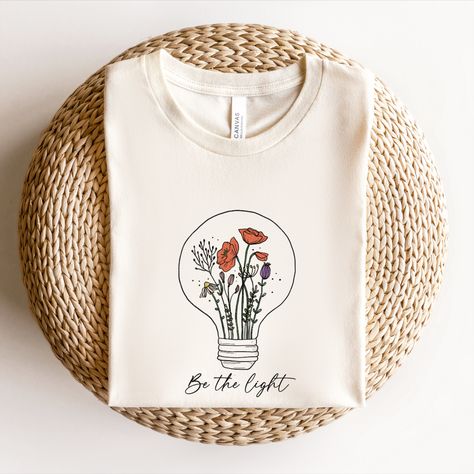 Floral shirts for women, Spring Shirt. This design with the quote "Be the light" and a lovely wildflower growing inside a light bulb is the perfect way to inspire yourself and others to shine bright and make a positive impact on the world. The beautiful illustration features a wildflower growing inside a light bulb, reminding you to nurture your own inner light and let it shine. The quote "Be the light" is a powerful reminder to be a positive influence and make a difference in the world. Roots Clothing, Light Flowers, Quote Graphic, Floral Shirts, Positive Influence, Be The Light, Inspiration Quote, Beautiful Illustration, Graphic Quotes