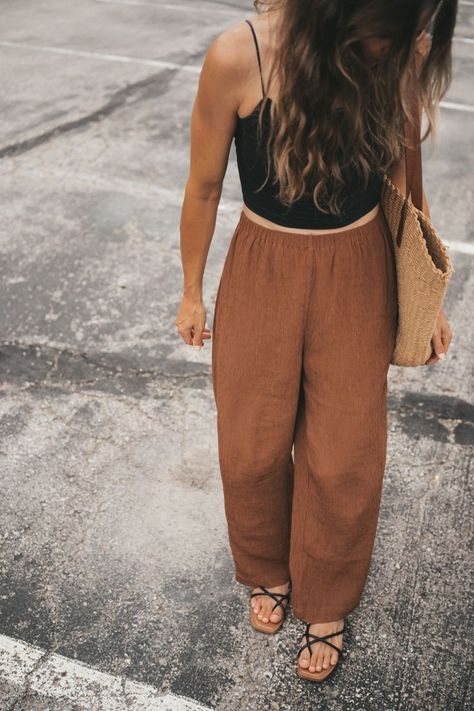 ashley wearing a black cropped cami, brown loose fitting tapered pants, black strappy sandals, and a woven tote bag walking through a parking lot Modern Boho Fashion Winter, Black And Brown Outfit Summer, Brown Linen Pants Outfit Summer, Boho Linen Outfit, Cool Summer Outfits Women, Brown Pants Outfit Summer, Minimalist Summer 2024 Outfits, Boho Minimalist Outfits, Olive Linen Pants Outfit