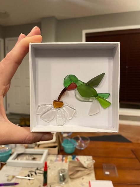 Hand holding up small shadow box. Inside shadow box are pieces of sea glass made in the shape of a daisy and a ruby throated hummingbird. Beach Glass Ideas, Sea Glass On Canvas, Sea Glass Hummingbird, Sea Glass Turtle Art, Brown Sea Glass Art, Sea Glass Canvas Art, Beach Glass Art Ideas, Sea Glass Resin Art, Sea Glass Animals