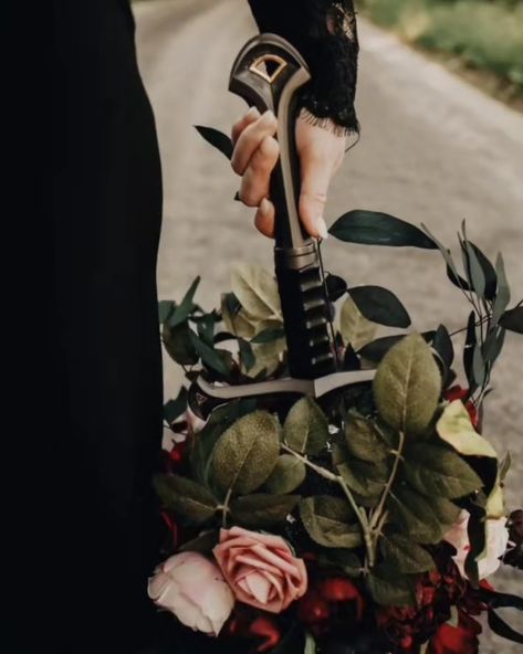 Bride in a black wedding dress holding a bouquet of pink and red flowers with lots of leaves downward by a sword hilt on a path. Gothic Wedding Theme, Lotr Wedding, Dark Wedding Theme, Forest Theme Wedding, Pagan Wedding, Viking Wedding, Enchanted Forest Wedding, Medieval Wedding, Goth Wedding